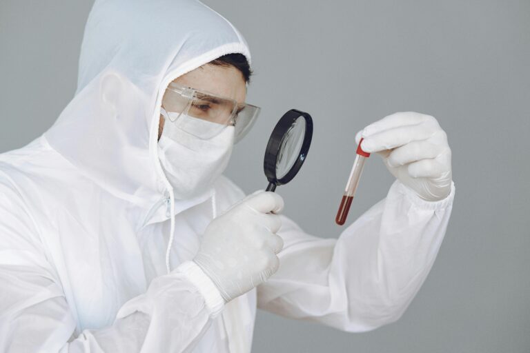A scientist in protective gear examines a blood sample through a magnifying glass.